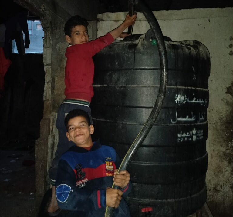 Portable Water Vehicle Delivers Water to Families in Gaza Children in Gaza standing in front of the water tank that is being filled with water from the vehicle.
